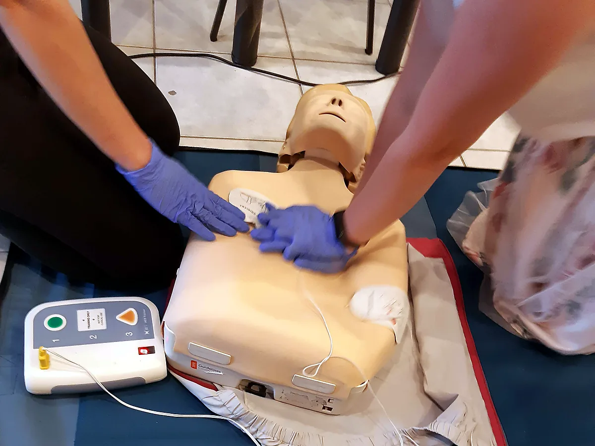 Szkolenie z obsługi AED: dwie osoby w rękawiczkach przy fantomie — jedna dociska elektrodę pod prawym obojczykiem, druga prowadzi uciśnięcia klatki piersiowej; obok trenażer AED z przyciskami 1-2-3