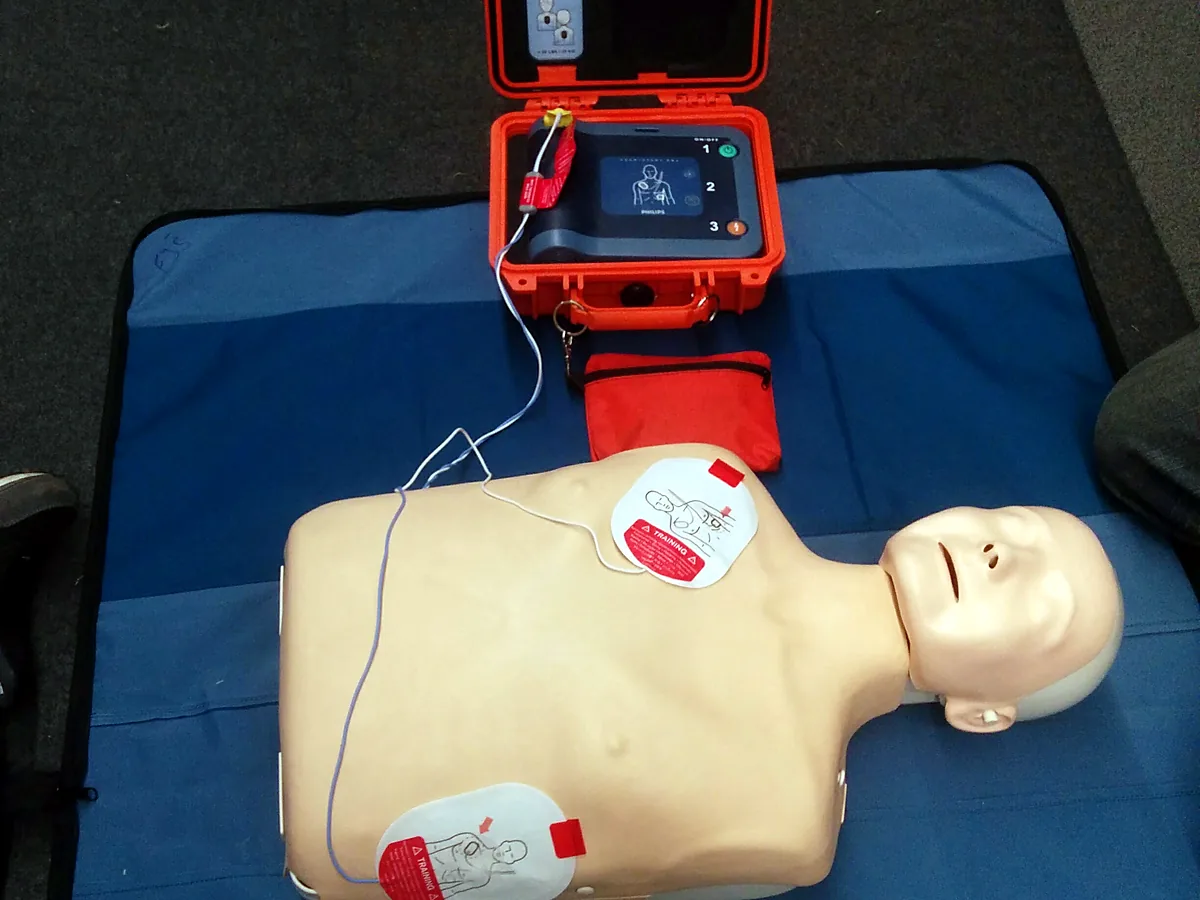Trenażer AED w pomarańczowej walizce transportowej podłączony do fantomu osoby dorosłej; elektroda naklejona pod prawym obojczykiem i druga po lewej stronie klatki piersiowej, w linii pachowej