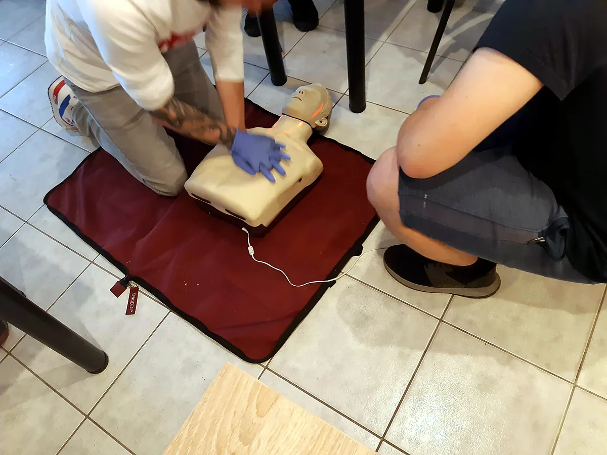 Uczestnik szkolenia prowadzi RKO na fantomie podłączonym do trenażera AED, obok w przysiadzie czeka druga osoba gotowa przejąć uciśnięcia