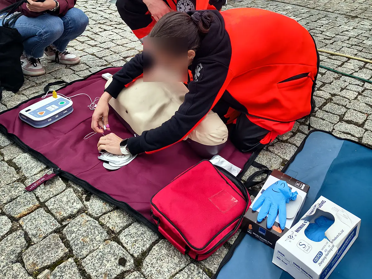 Szkolenie z AED w plenerze na kostce brukowej: ratowniczka w kurtce ratownictwa medycznego nakleja elektrody treningowe na fantomie, obok trenażer AED, czerwona torba medyczna i pudełka rękawiczek; twarz zamazana